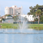 A body of water with a small fountain spouting water out in upward dispersal. Green grass and trees surround the water with a metal sculpture on the right and white and gray buildings directly behind the spouting water.