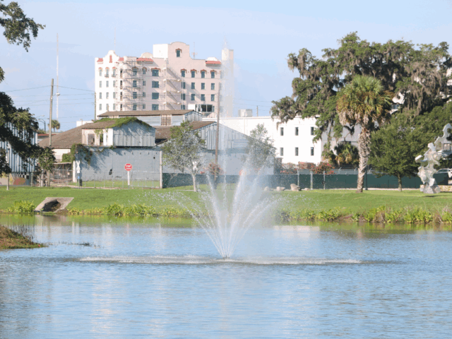 A body of water with a small fountain spouting water out in upward dispersal. Green grass and trees surround the water with a metal sculpture on the right and white and gray buildings directly behind the spouting water.