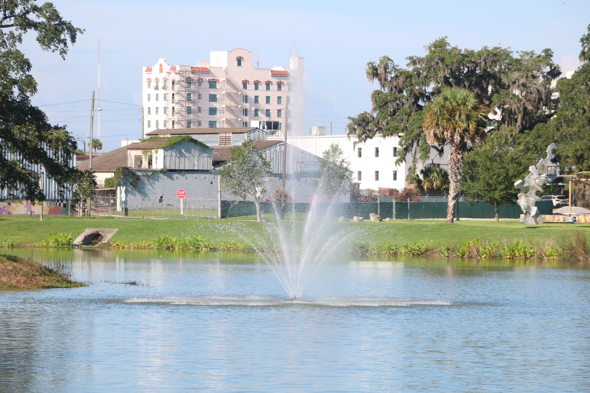 A body of water with a small fountain spouting water out in upward dispersal. Green grass and trees surround the water with a metal sculpture on the right and white and gray buildings directly behind the spouting water.