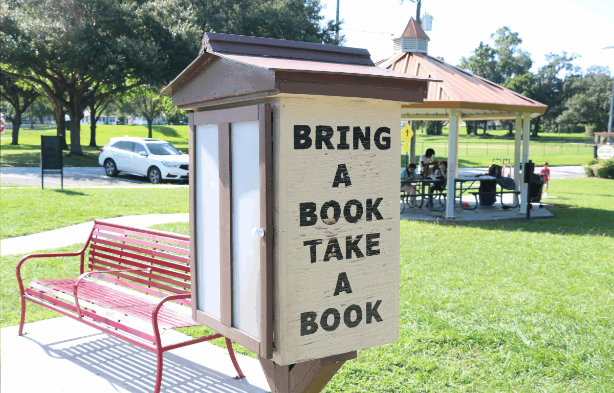 A small wooden free library box in a park with the words "BRING A BOOK TAKE A BOOK" painted on the side. A red bench sits nearby, and a covered picnic area with people is visible in the background. Trees and a white car are in the distance.