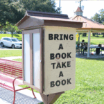 A small wooden free library box in a park with the words "BRING A BOOK TAKE A BOOK" painted on the side. A red bench sits nearby, and a covered picnic area with people is visible in the background. Trees and a white car are in the distance.