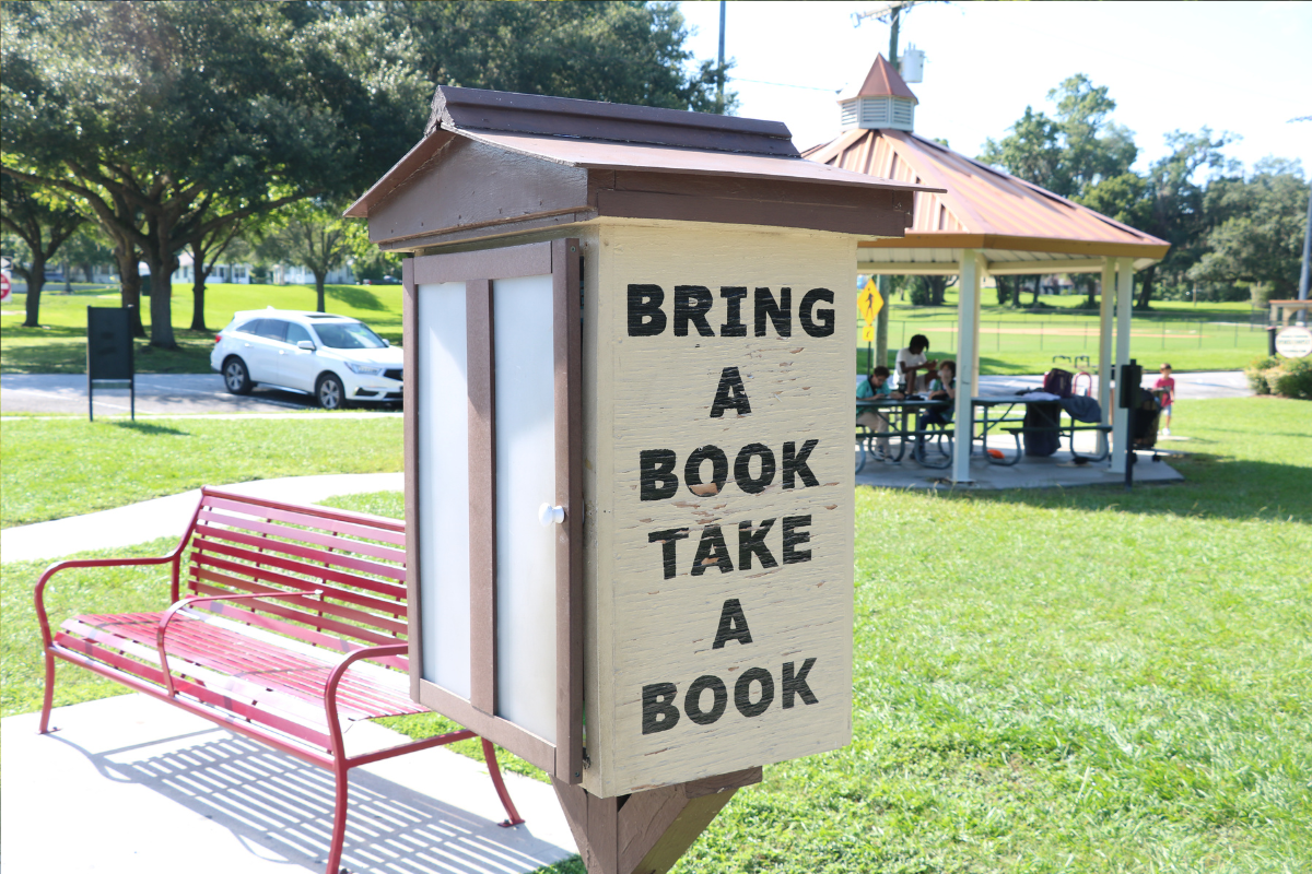 A small wooden free library box in a park with the words "BRING A BOOK TAKE A BOOK" painted on the side. A red bench sits nearby, and a covered picnic area with people is visible in the background. Trees and a white car are in the distance.