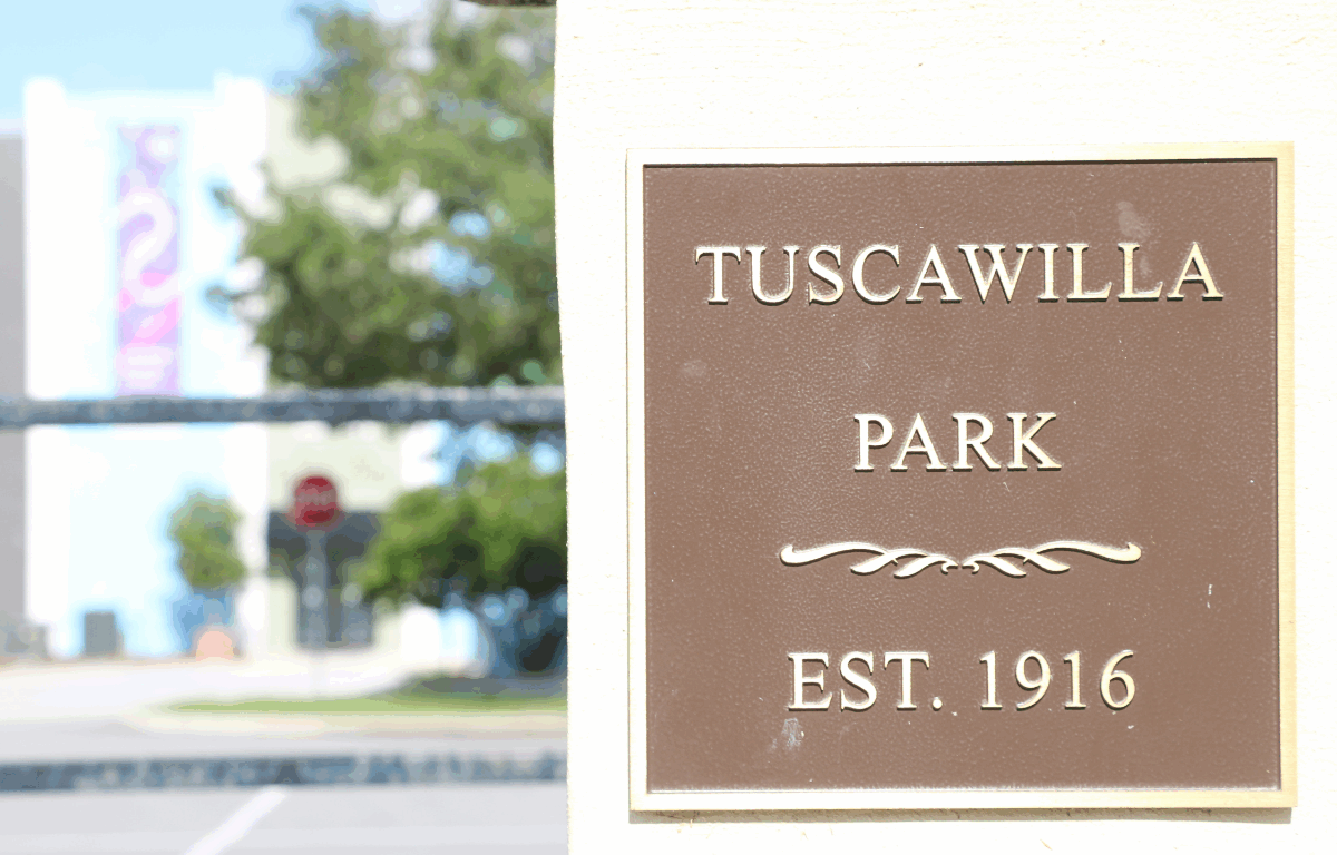A close-up of a brown plaque mounted on a light-colored wall reads "Tuscawilla Park, Est. 1916" in gold lettering, with a decorative flourish in the middle. A blurred background shows a street, trees, and a building.