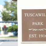A close-up of a brown plaque mounted on a light-colored wall reads "Tuscawilla Park, Est. 1916" in gold lettering, with a decorative flourish in the middle. A blurred background shows a street, trees, and a building.