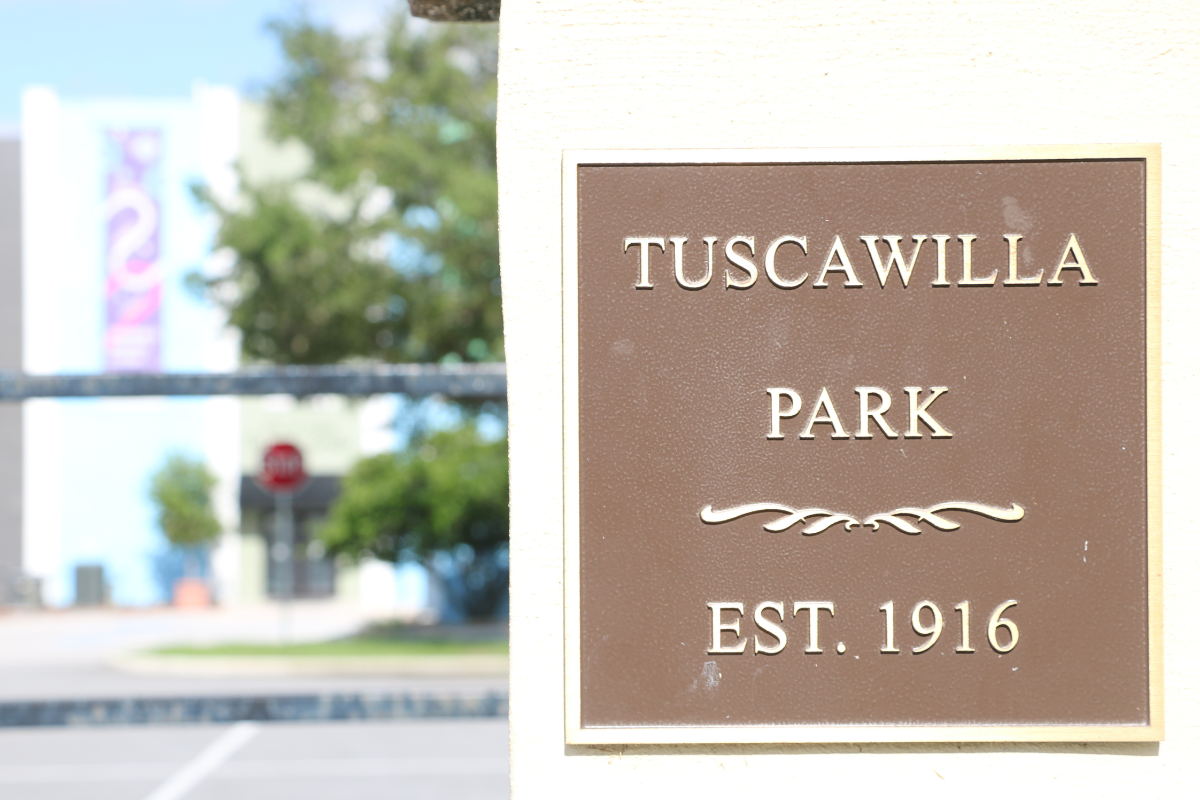 A close-up of a brown plaque mounted on a light-colored wall reads "Tuscawilla Park, Est. 1916" in gold lettering, with a decorative flourish in the middle. A blurred background shows a street, trees, and a building.