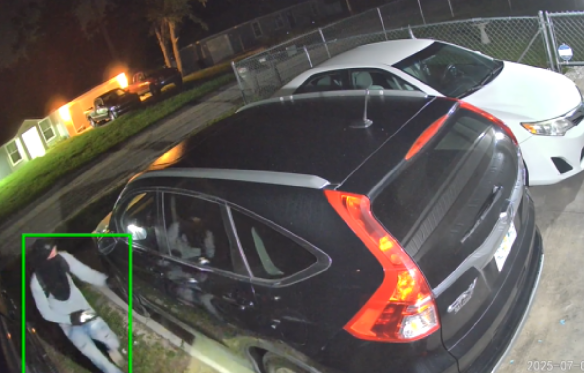 A nighttime security camera image shows a person wearing a hood, mask, and light-colored clothing approaching a parked black SUV in a residential driveway. The individual appears to be holding a flashlight. Nearby are other parked vehicles, a fenced yard, and illuminated houses in the background. The timestamp on the image reads "2025-07-21.