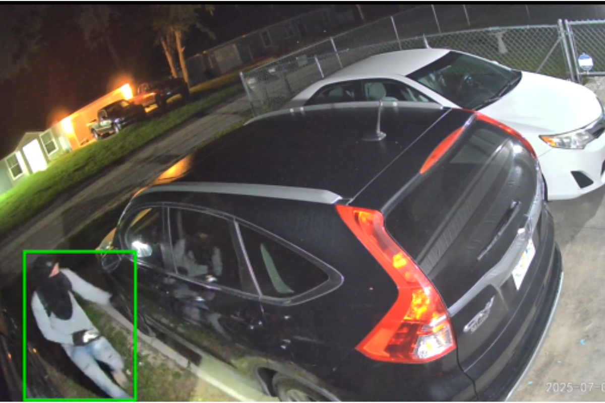 A nighttime security camera image shows a person wearing a hood, mask, and light-colored clothing approaching a parked black SUV in a residential driveway. The individual appears to be holding a flashlight. Nearby are other parked vehicles, a fenced yard, and illuminated houses in the background. The timestamp on the image reads "2025-07-21.