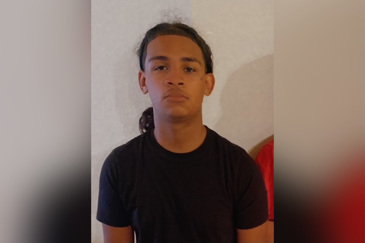 A teenage boy with medium brown skin and long dark hair pulled back, wearing a black t-shirt, stands in front of a neutral-colored wall. He has a serious expression on his face.