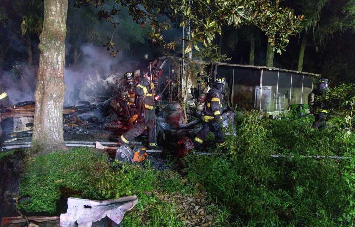 Firefighters in a wooden area fighting a house fire in a single-wide mobile home.