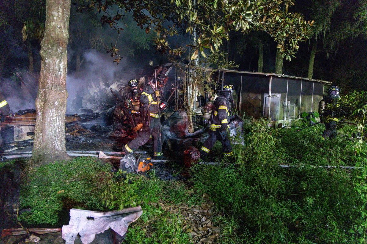 Firefighters in a wooden area fighting a house fire in a single-wide mobile home.