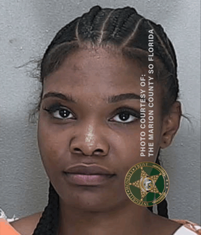 Mugshot of a woman with braided hair pulled back, wearing an orange jail uniform, staring straight at the camera.