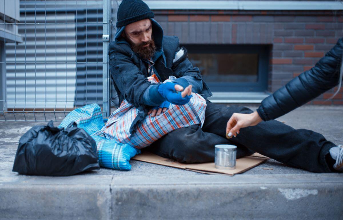 Man experiencing homelessness sitting on sidewalk receiving money from passerby.