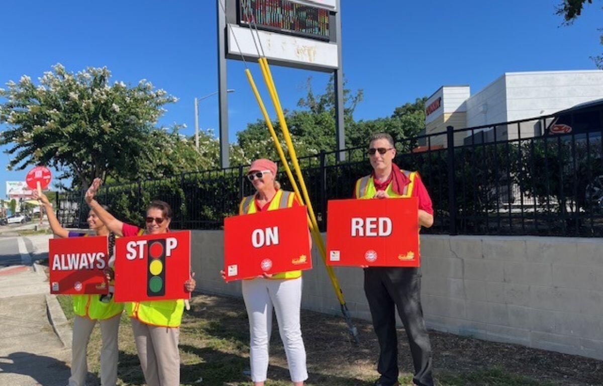 To the extreme left, a woman holding a fan and a placard, four people in safety vests holding placard with writing and images, with a sidewalk in the foreground and wall with metal fencing top, with a sign behind the people, retail space and trees in the background.