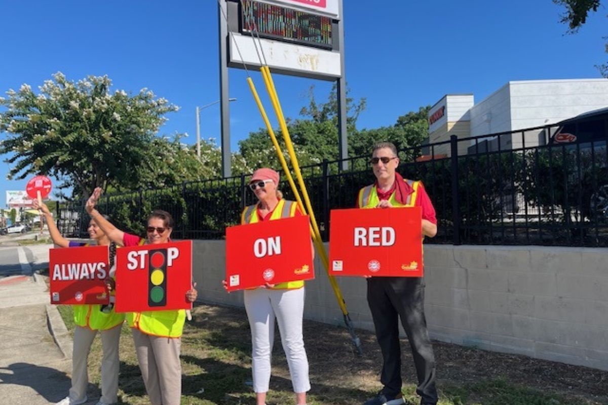 To the extreme left, a woman holding a fan and a placard, four people in safety vests holding placard with writing and images, with a sidewalk in the foreground and wall with metal fencing top, with a sign behind the people, retail space and trees in the background.