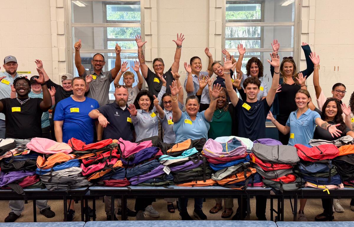 A group of people in different colored shirts and yellow nametags stand behind folded tables filled with colored backpacks; their arms raised in the air.