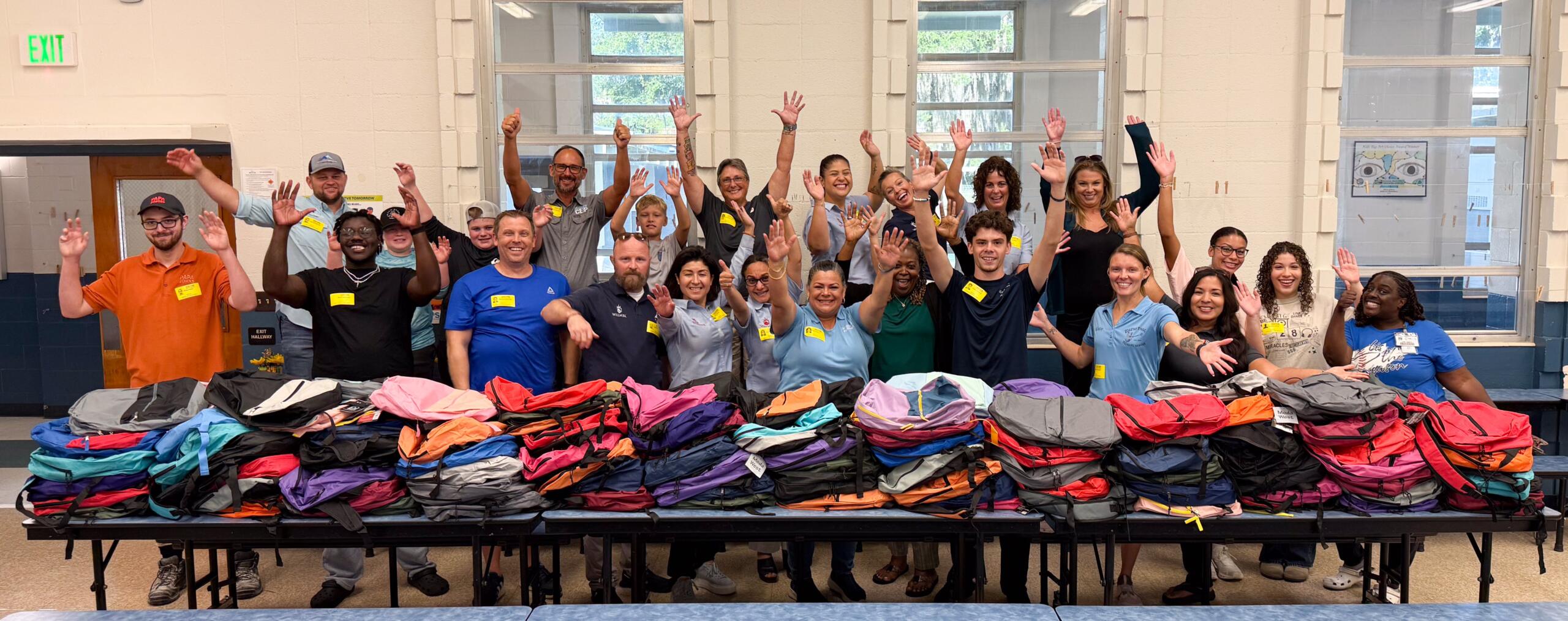 A group of people in different colored shirts and yellow nametags stand behind folded tables filled with colored backpacks; their arms raised in the air.