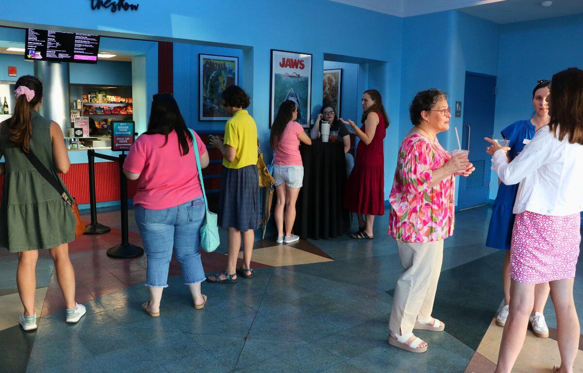 A room of guests dressed in pink and Barbie-themed outfits pose in front of a decorated backdrop at a Barbie-themed event.