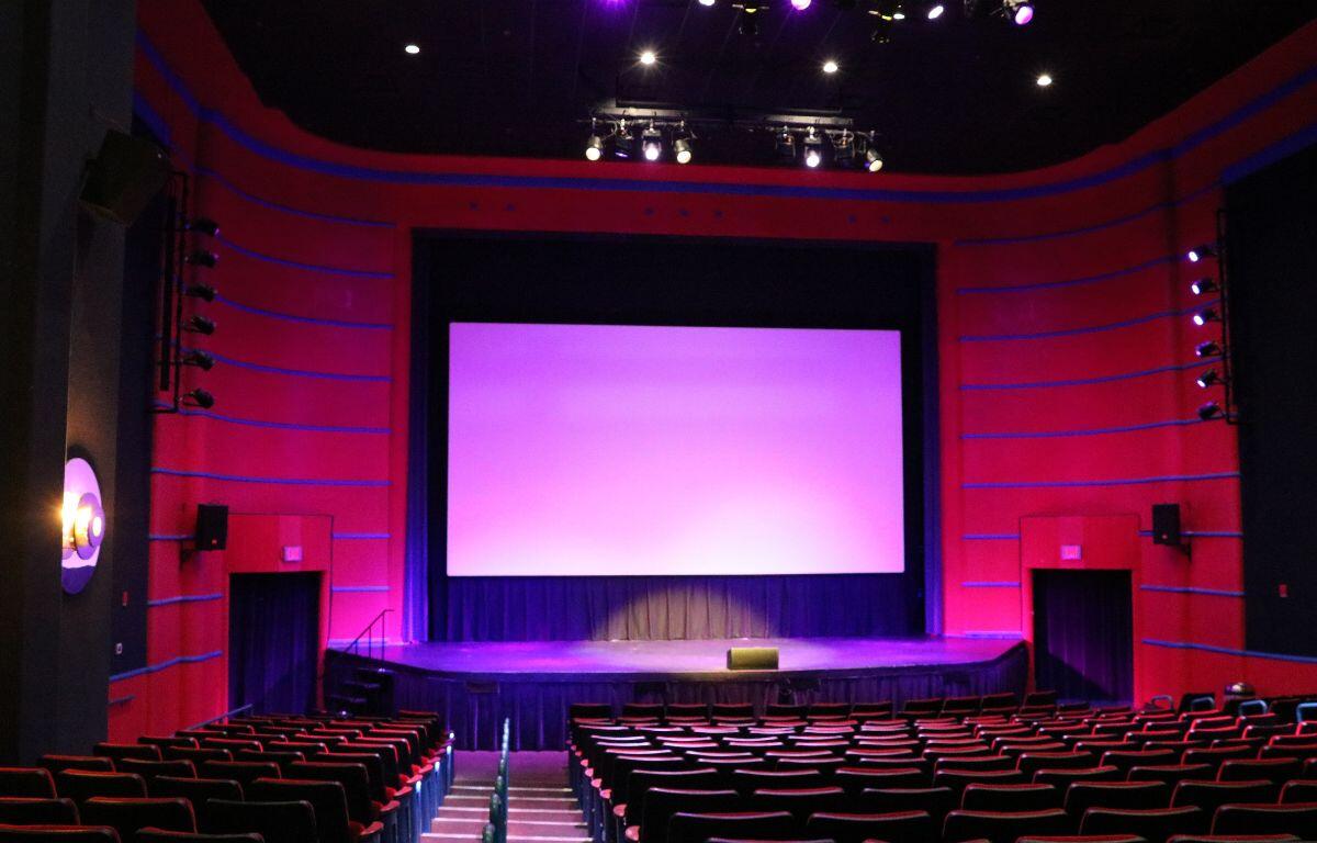A theatre with multiple rows of red seats and a large screen is dimly lit with pink and magenta stage lights. The theatre is red with blue stripes and a large stage just below the screen.