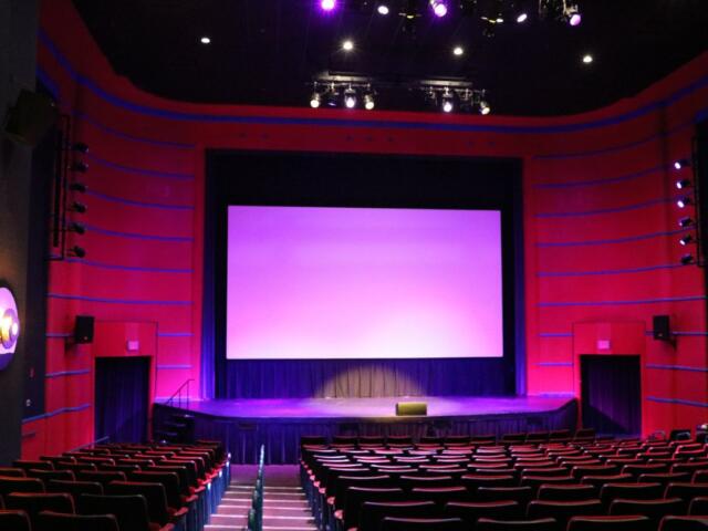 A theatre with multiple rows of red seats and a large screen is dimly lit with pink and magenta stage lights. The theatre is red with blue stripes and a large stage just below the screen.