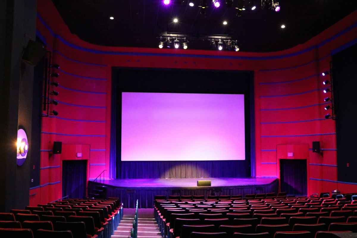 A theatre with multiple rows of red seats and a large screen is dimly lit with pink and magenta stage lights. The theatre is red with blue stripes and a large stage just below the screen.