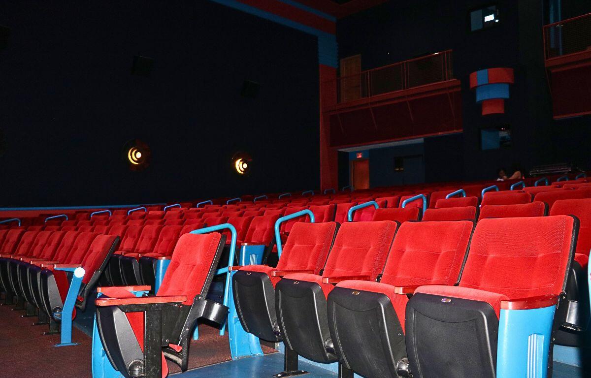 Bright red seating in rows fills the isles of a movie theatre with a dimly lit background as two guests wait for a movie to begin in the dim background.