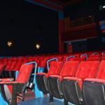 Bright red seating in rows fills the isles of a movie theatre with a dimly lit background as two guests wait for a movie to begin in the dim background.