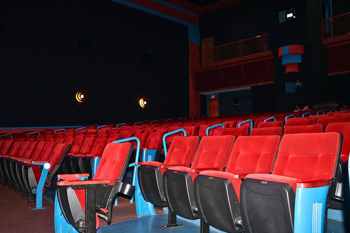 Bright red seating in rows fills the isles of a movie theatre with a dimly lit background as two guests wait for a movie to begin in the dim background.