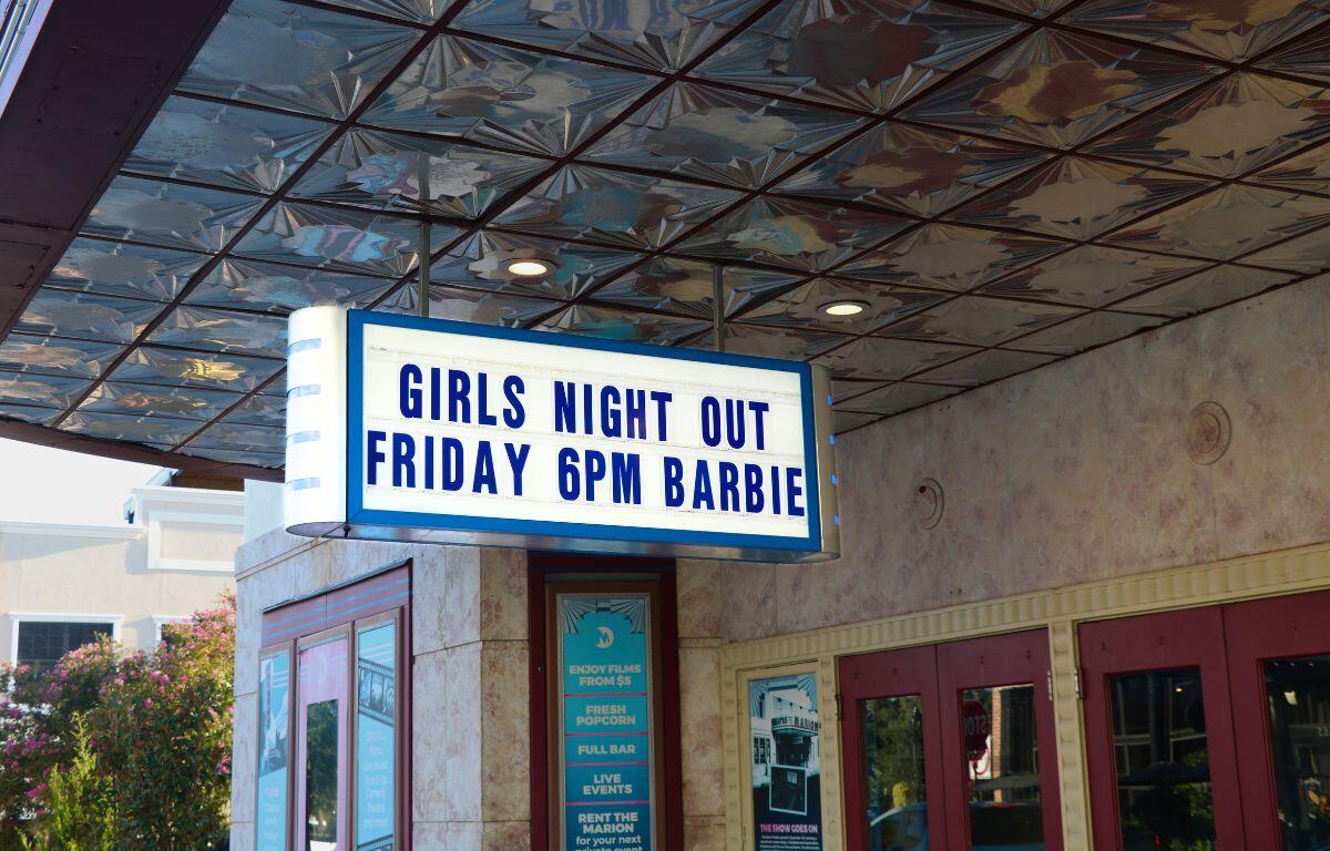 A white, hanging sign with blue accents reads, "Girls Night Out" on the top line and "Friday 6 p.m. Barbie" on the bottom. The sign hangs outside of a theatre with signs advertising the theatre with red double doors toward the right leading into the lobby.