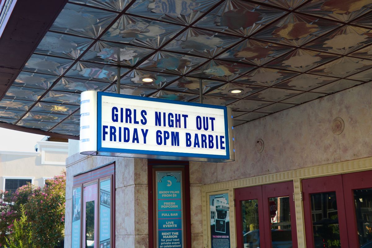 A white, hanging sign with blue accents reads, "Girls Night Out" on the top line and "Friday 6 p.m. Barbie" on the bottom. The sign hangs outside of a theatre with signs advertising the theatre with red double doors toward the right leading into the lobby.