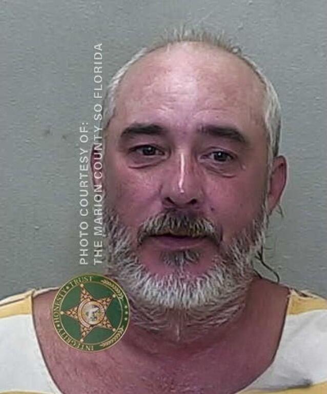 Mugshot of a bald man with a short gray beard, wearing a white and orange prison uniform, against a gray background.