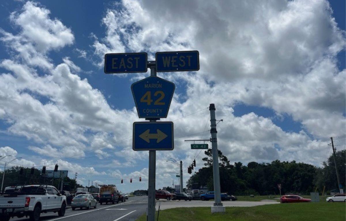 To the left a stretch of roadway with cars, in the background white clouds, blue skies, a traffic sign, and in the far distance a traffic signal, to the right in the background a tree line. The focal point is signs, with two rectangular signs with writing on top of a blue pentagon shaped sign with writing. with a rectangular sign underneath with letters.