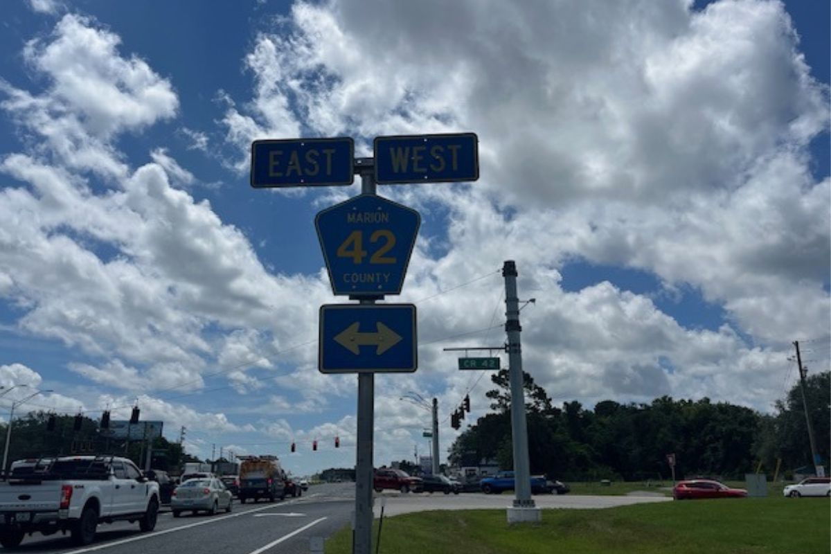 To the left a stretch of roadway with cars, in the background white clouds, blue skies, a traffic sign, and in the far distance a traffic signal, to the right in the background a tree line. The focal point is signs, with two rectangular signs with writing on top of a blue pentagon shaped sign with writing. with a rectangular sign underneath with letters.