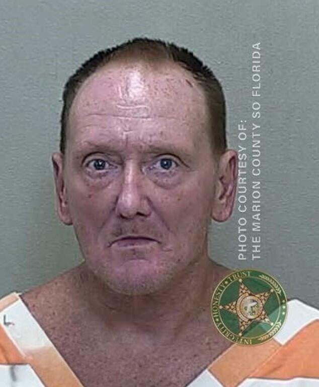Mugshot of a man with thinning hair, wearing a white and orange prison uniform, against a gray background.