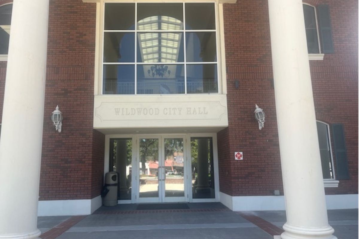 Extreme left, brick wall in background, in the middle the interior of the lobby is visible through the window, facade in the background over the window treatment, which appears to be limestone, stenciled lettering, deeply inset in the background glass windows and glass doors, to the right brick facade, small sign with an insignia, to the left and right of the glass and metal sconce lighting fixtures, to the right another column and more brick. A concrete walkway is in the foreground leading to the doors that open to the vestibule.