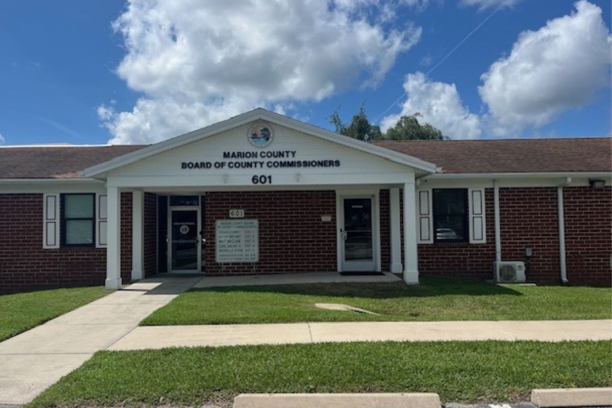Background is a blue sky, white clouds, shade trees behind the roof of the building, middle ground, a building with white columns, inset with window and doors and brick, in front of the building, sod and sidewalks, and in the foreground, a parking lot.