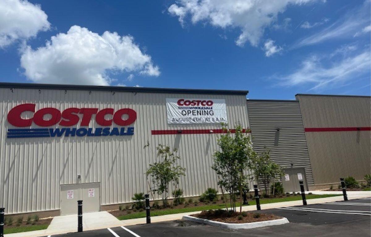 A large warehouse with lettering on it and a banner with lettering on it hanging down in the middle of the photo, in the middle of the photo there's flora and a sidewalk, and in the foreground a parking lot.