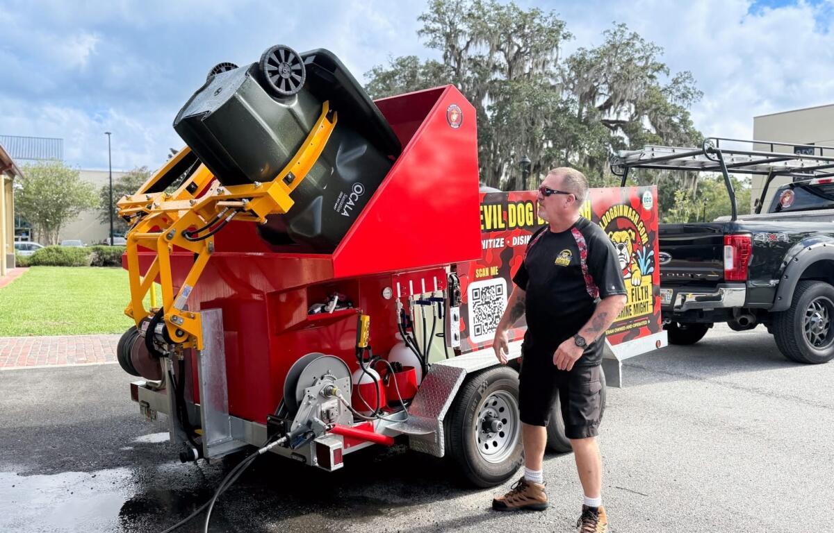 A man wearing sunglasses and work boots stands beside a bright red Devil Dog Bin Wash truck, watching as a green trash bin is lifted into the machine’s yellow hydraulic arms for cleaning.