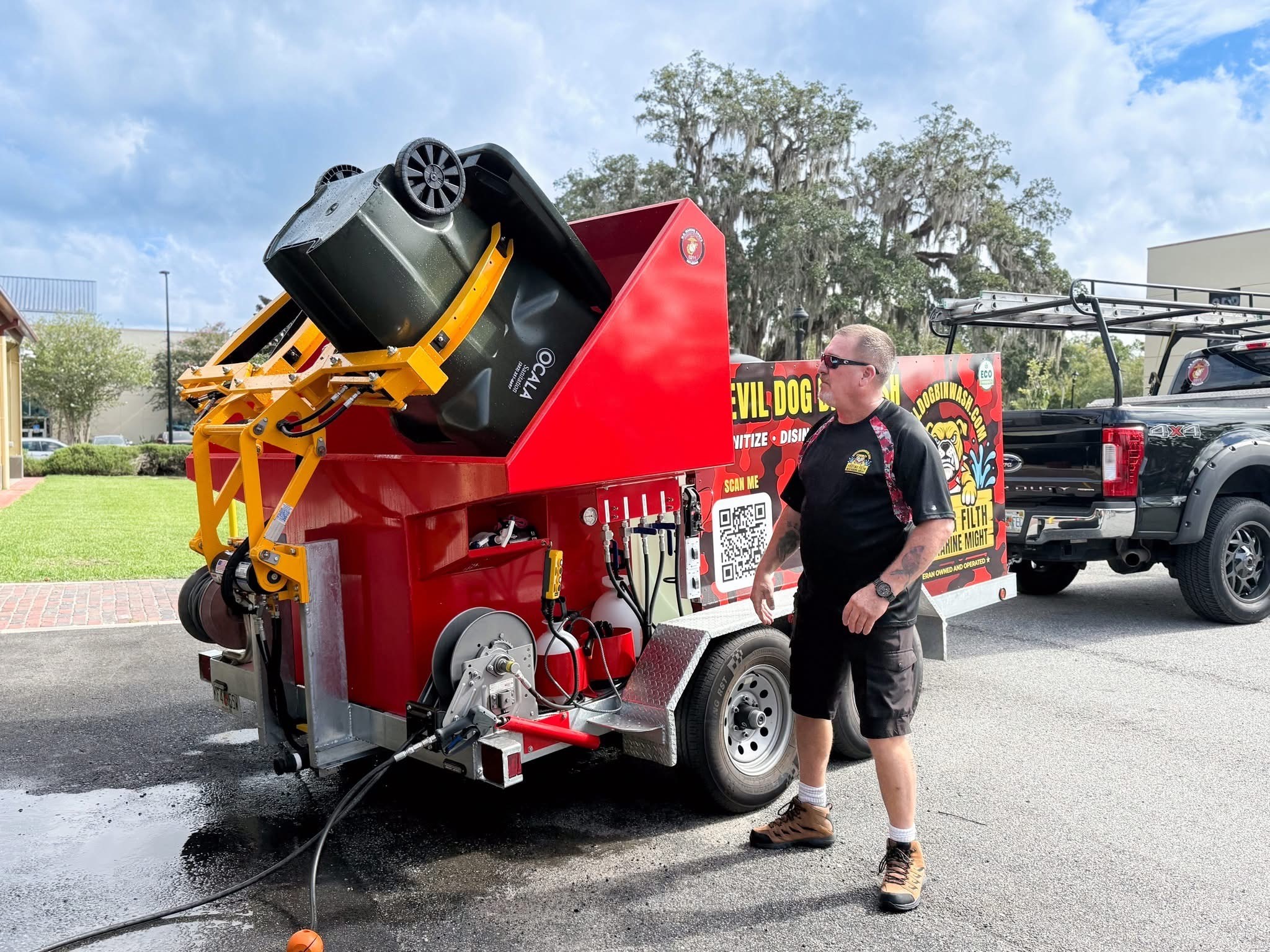 A man wearing sunglasses and work boots stands beside a bright red Devil Dog Bin Wash truck, watching as a green trash bin is lifted into the machine’s yellow hydraulic arms for cleaning.