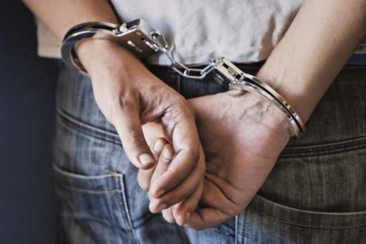 A close-up photo of a person’s hands cuffed together with metal handcuffs, resting on their lap.