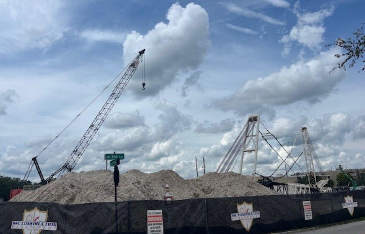 In the background, blue skies, white clouds, in the center, cranes and piles of dirt, in the foreground a construction fence with logos on it and a street sign.