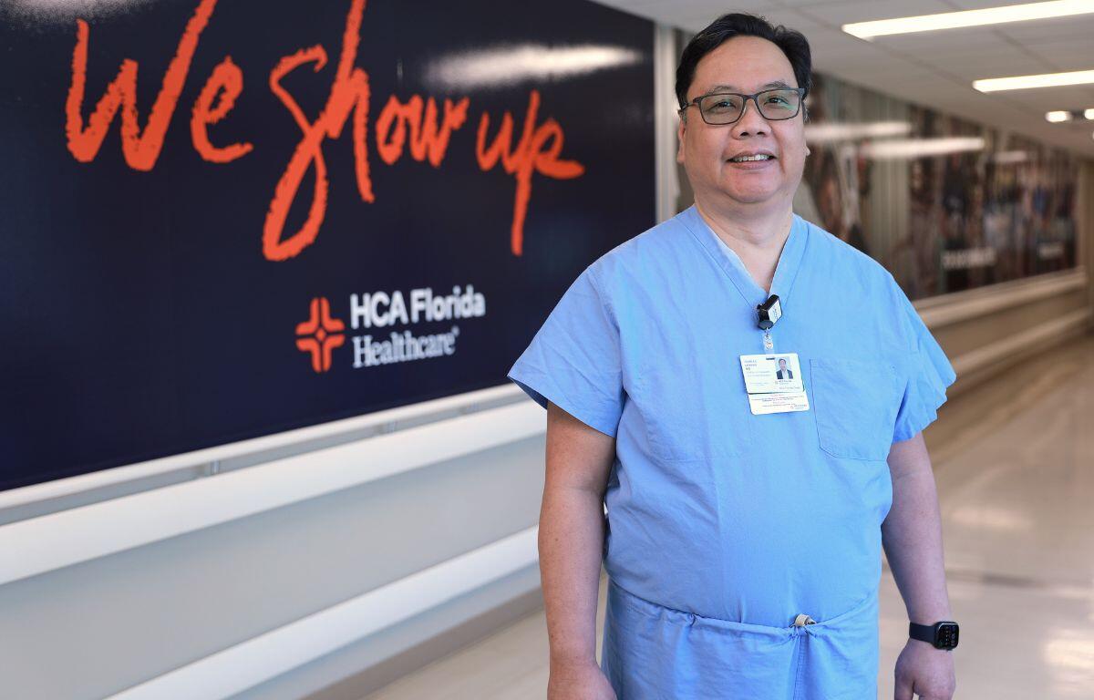 In the foreground a man wearing glasses and blue scrubs, wearing an identification badge. the background has a black wall with red and white lettering, and below that the wall is white, with a long hallway to the right with photos on it.