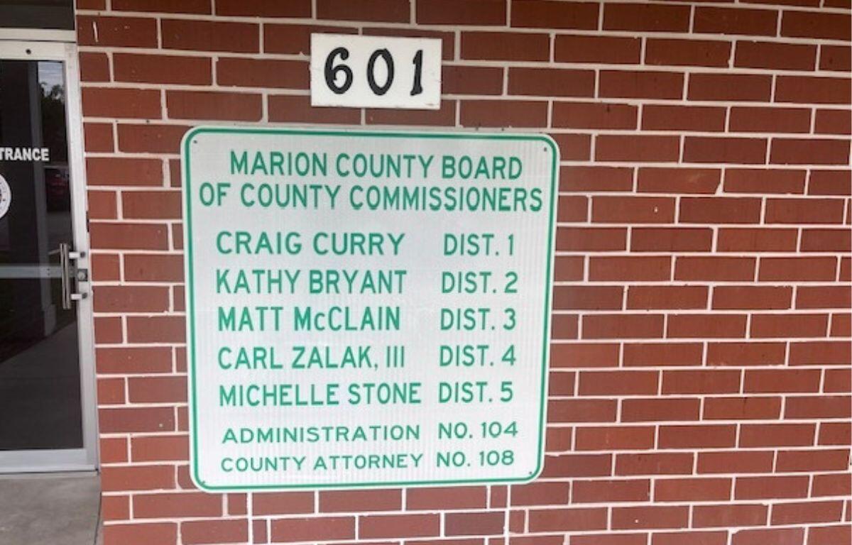A door to the extreme left, a white placard with names and numbers, with a smaller white placard with numbers, affixed to a brick wall.
