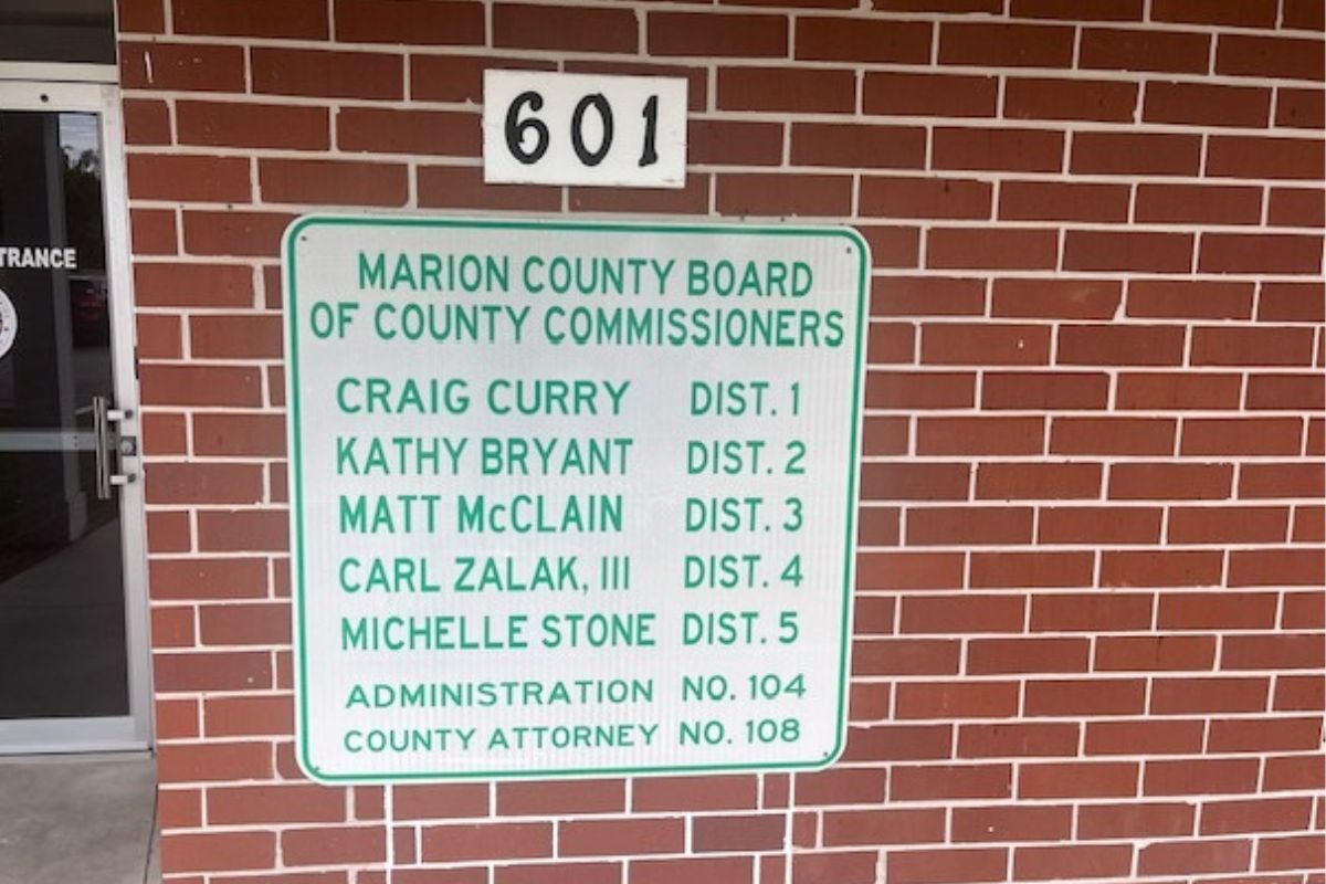 A door to the extreme left, a white placard with names and numbers, with a smaller white placard with numbers, affixed to a brick wall.