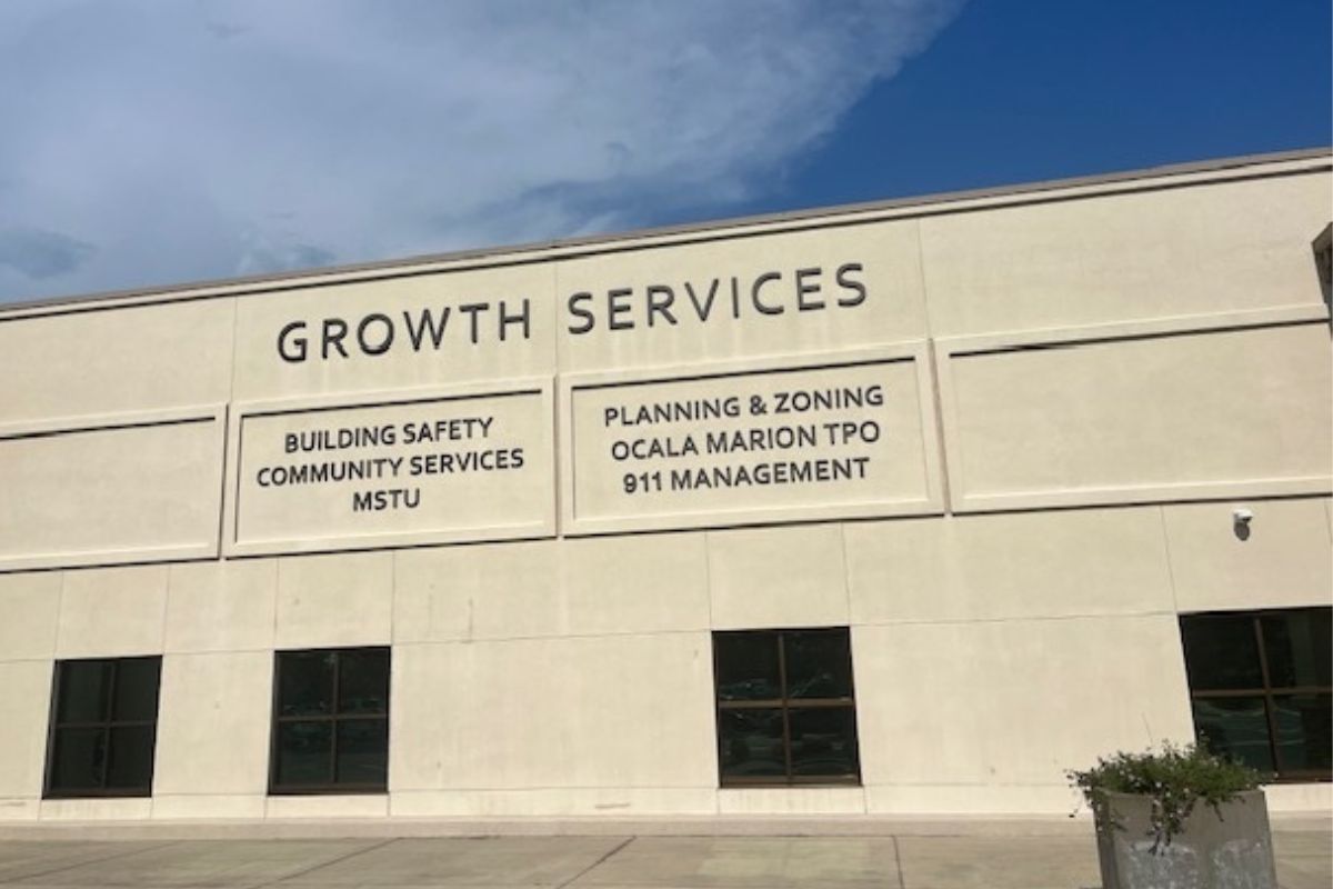Background has a blue sky and white clouds, over a beige colored building, with lettering and windows, with ornamental plans in planter and a parking lot in the foreground.