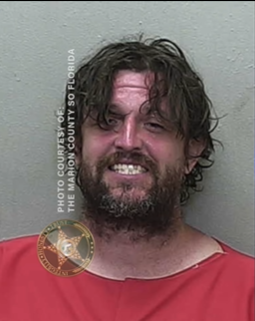 Mugshot of Eric Johnson, a man with messy dark hair covering part of his face, a scruffy beard, and a slight smile, wearing a red jail uniform against a gray background.