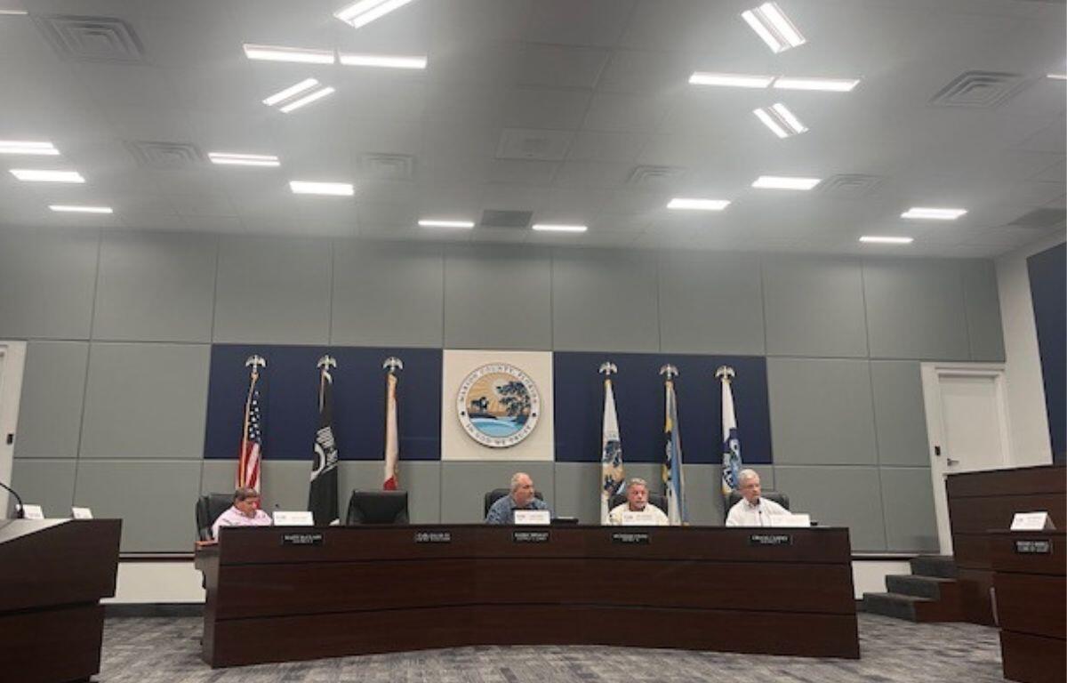 Four men sitting behind a wood dais, with carpeting in the foreground, three flags on each side of a large insignia with an image and lettering in the background.