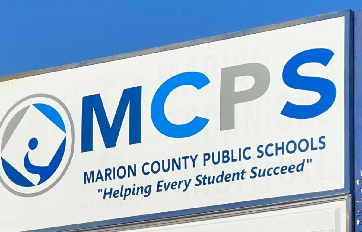 A blue sky in the background, a white sign with an insignia to the left and lettering on it.