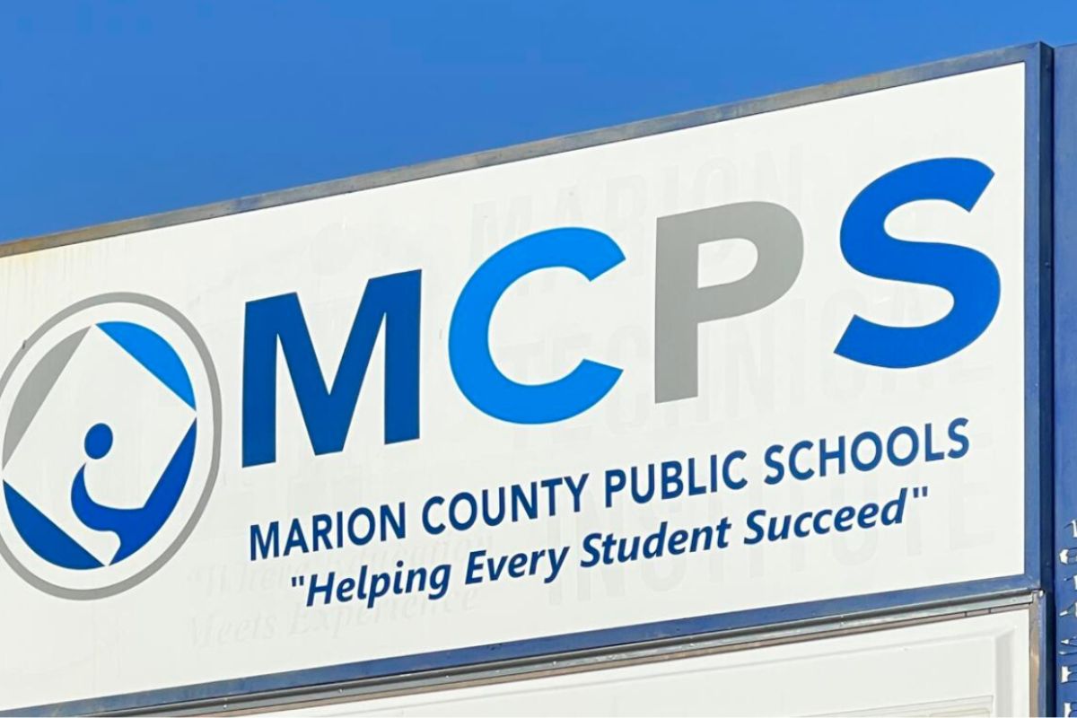 A blue sky in the background, a white sign with an insignia to the left and lettering on it.