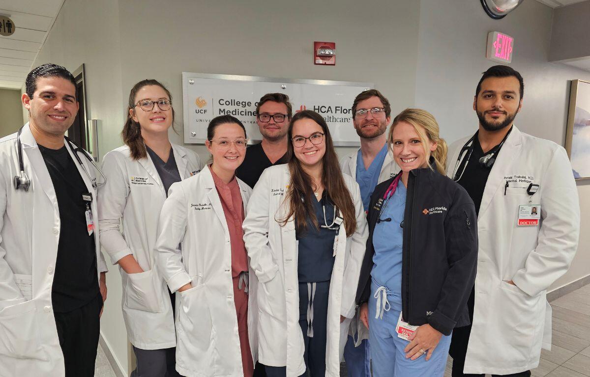 A group of men and women wearing doctors smocks and scrubs standing against a wall.