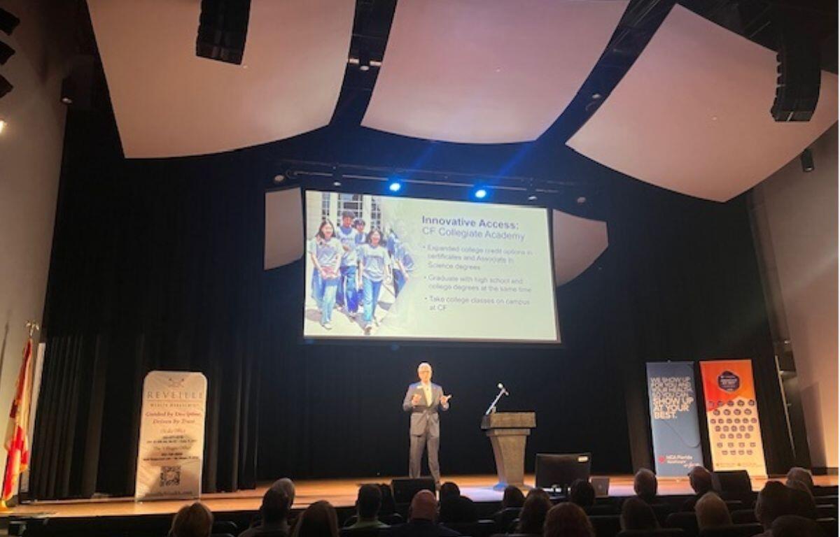 A black background, with flags, a large placard with writing to the left, in the center of the stage man, with a screen over his head, and a podium and hanging banners to his right, In the foreground there are auditorium seats and a crowd in their seats.
